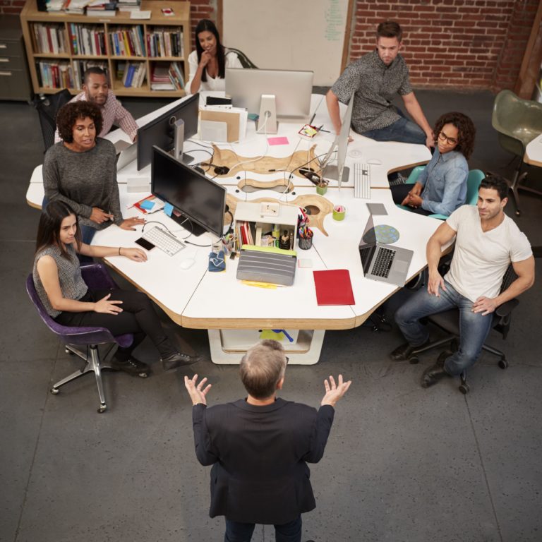 Business team around a table, listening to a man who is standing, speak